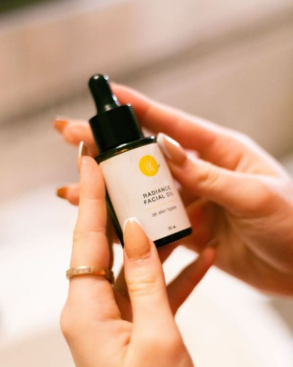 Two white hands with manicured nails hold a bottle of Radiance Facial Oil